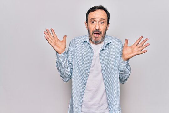 Middle age handsome man wearing casual denim shirt standing over isolated white background clueless and confused with open arms, no idea and doubtful face.