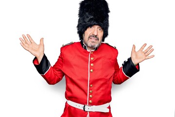 Middle age handsome wales guard man wearing traditional uniform over white background clueless and confused expression with arms and hands raised. Doubt concept.