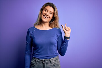 Young beautiful blonde woman wearing casual t-shirt over isolated purple background with a big smile on face, pointing with hand finger to the side looking at the camera.