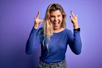 Young beautiful blonde woman wearing casual t-shirt over isolated purple background shouting with crazy expression doing rock symbol with hands up. Music star. Heavy music concept.