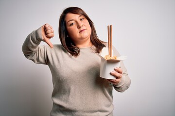 Beautiful plus size woman eating asian noodles food from box over isolated background with angry...