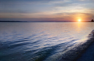 Fototapeta premium Purple sunset over a large pond. Calm water in the river. Silhouettes of trees on the horizon. The last rays of the sun are reflected in the water. Light clouds in the sky. European nature.