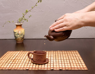 Woman is pouring tea from a teapot into a cup. Chinese tea ceremony. Vase with a branch in the background in blur.