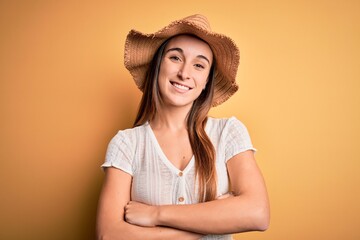 Young beautiful woman wearing casual t-shirt and summer hat over isolated yellow background happy face smiling with crossed arms looking at the camera. Positive person.