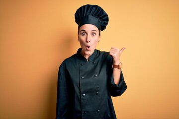 Young beautiful chef woman wearing cooker uniform and hat standing over yellow background Surprised pointing with hand finger to the side, open mouth amazed expression.