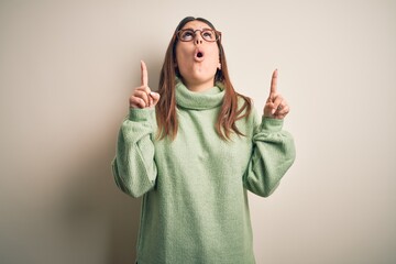 Young beautiful woman wearing casual sweater standing over isolated white background amazed and surprised looking up and pointing with fingers and raised arms.