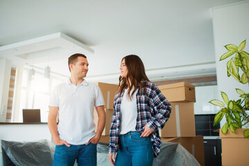 A young married couple in the living room in the house stand near unpacked boxes. hey are happy about new home. Moving, buying a house, apartment concept.
