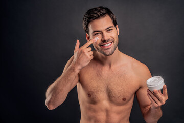 Handsome young man isolated. Portrait of shirtless muscular man is standing on grey background and using face cream, smiling and looking at camera. Men care concept.