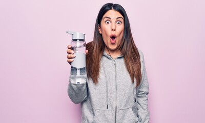 Young beautiful brunette sporty woman drinking bottle of water over isolated pink background scared and amazed with open mouth for surprise, disbelief face