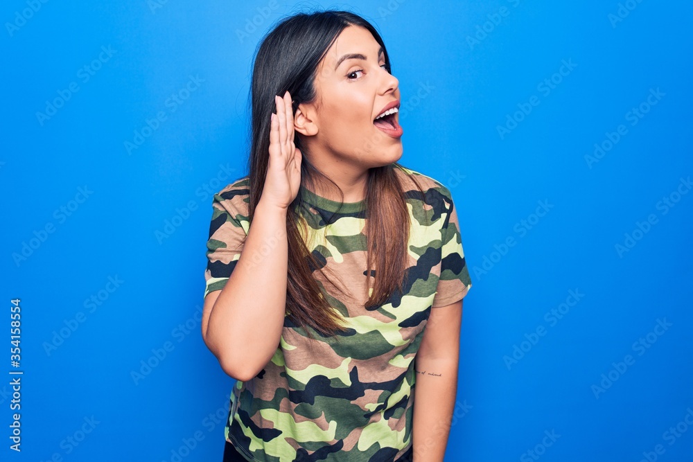 Sticker Young beautiful brunette woman wearing camouflage military t-shirt over blue background smiling with hand over ear listening and hearing to rumor or gossip. Deafness concept.