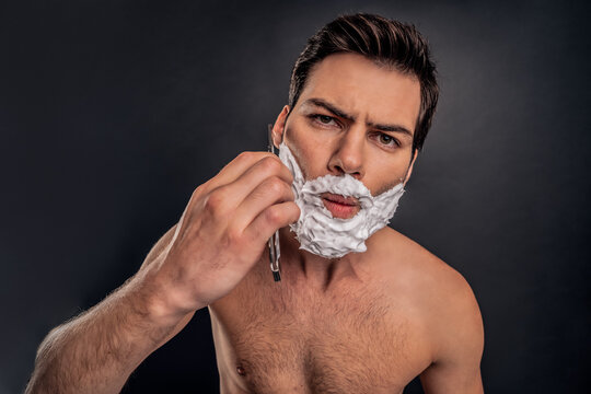 Young handsome man with shaving cream on his face, grooming his beard with straight razor and looking at camera