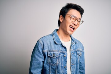 Young handsome chinese man wearing denim jacket and glasses over white background winking looking...