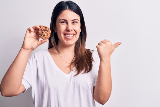 Young beautiful brunette woman eating chocolate cookie over isolated white background pointing thumb up to the side smiling happy with open mouth