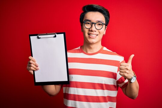 Young Handsome Chinese Inspector Man Wearing Glasses Holding Clipboard Checklist Happy With Big Smile Doing Ok Sign, Thumb Up With Fingers, Excellent Sign