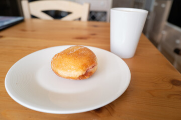 A jam doughnut on a white plate next to a white mug cup on a wood kitchen table with a wooden chair. Soft focus