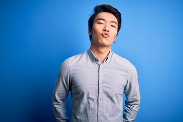 Fototapeta premium Young handsome chinese man wearing casual shirt standing over isolated blue background looking at the camera blowing a kiss on air being lovely and sexy. Love expression.