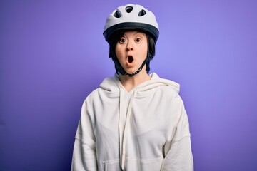 Young down syndrome cyclist woman wearing security bike helmet over purple background afraid and shocked with surprise expression, fear and excited face.