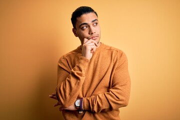 Young brazilian man wearing casual sweater standing over isolated yellow background with hand on chin thinking about question, pensive expression. Smiling with thoughtful face. Doubt concept.