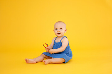 Funny little girl dressed in blue dress sitting and playing with pineapple isolated on yellow background. Summer holidays concept