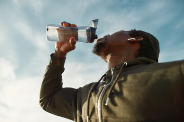 Taking a break. Young african male runner in sportswear drinking fresh water from a bottle while...