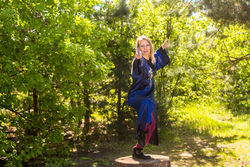 A woman in a blue kimono poses with a sword in the forest standing on a stump