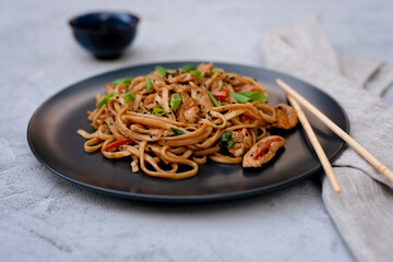 Asian udon noodles with chicken vegetables and teriyaki sauce with chopsticks and a linen napkin on a gray concrete background. Chinese and Japanese cuisine.