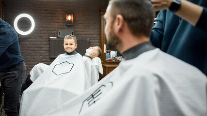 Family visiting barbershop. Cute and happy boy looking at his father with smile while getting...