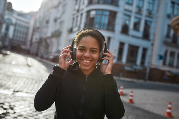 Fototapeta premium Best jogging music. Happy african female runner wearing headphones looking at camera and smiling. Young woman in sportswear running outdoors. Morning jog