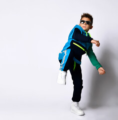Little kid in sunglasses, colorful sport suit and sneakers. He performing a kick by his leg, posing isolated on white