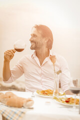 Smiling caucasian man holds glass of red wine sitting at table on the coast outdoors and looking at side. Romantic date concept. Celebratory toast concept. Toned image.