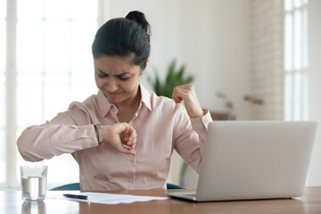 Stressed young indian businesswoman sitting in office, looking in hurry at wristwatch, irritated by...