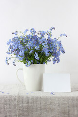blue forget-me-nots in a white mug and an empty paper card