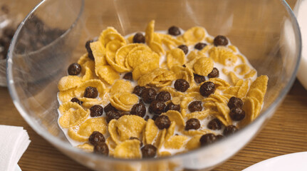 Glass bowl of Cornflakes with chocolate balls and milk on a wooden table. Cereal breakfast. Healthy quick food concept. Close up.