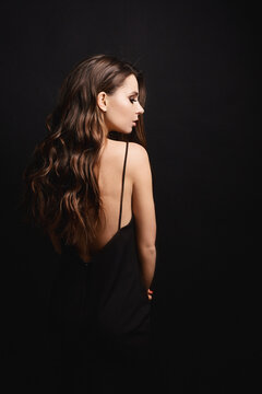 Brunette model girl in the black dress with nude back posing with her back at black background. Beauty and fashion concept