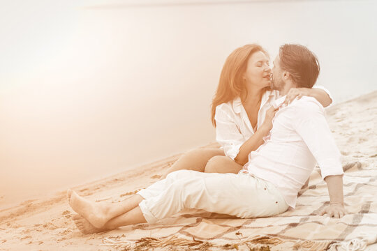 Couple in love gently hugs and kisses sitting on blanket in the sand near water. Mature couple in white in white clothes spend time together relaxing outdoors. Romantic date concept. Toned image.