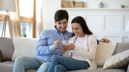 Happy young caucasian couple sit relax on sofa in living room using modern smartphone gadget shopping online together, smiling man and woman rest on couch at home, browse internet on cellphone