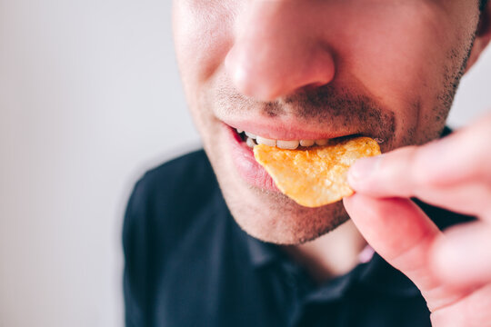 Young Man Isolated Over Background. Cut View And Close Up Of Guy Biting Piece Of Potato Chips With Teeth. Holding In It Hand. Tasty Delicious Fast Food.