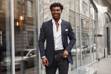 Black man in a city. Guy by the gray building. Man in a black suit.