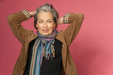 Graying mature woman making hairstyle correcting her hair by hands. creative woman gathered hair on the back of her head posing on a pink background.