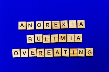 words anorexia, bulimia, overeating. Wooden blocks with an inscription on top on a blue background. Social problems of teenagers