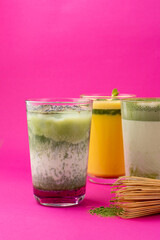 Close up of assortment of iced matcha lattes on minimalist magenta background, three glasses with traditional whisk