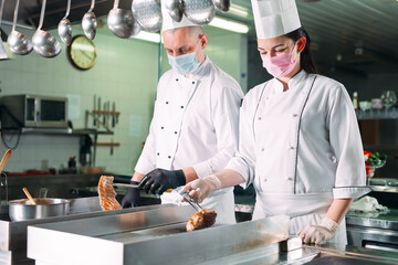 Chefs in protective masks and gloves prepare food in the kitchen of a restaurant or hotel.