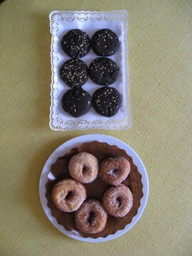 seis galletas de harina de algarroba cubiertas de frutos secos en bandeja con papel de blonda blanco para reposter&iacute;a y cinco rosquillas de an&iacute;s cubiertas de az&uacute;car en plato blanco con papel marr&oacute;n. Du