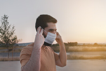 Young man with beard putting on a white face mask to protect himself of coronavirus pandemic. New normality