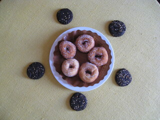 cinco rosquillas de anís, fritas, cubiertas de azúcar en plato blanco con papel marrón, rodeadas de cinco galletas de algarroba con frutos secos sobre mantel. Dulces artesanos, caseros, ecológicos.