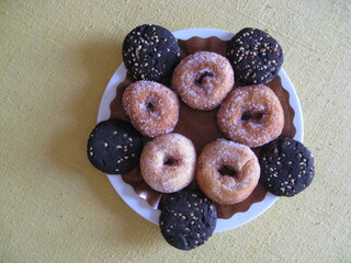 cinco rosquillas de anís, fritas, cubiertas de azúcar en plato blanco con papel marrón, rodeadas de cinco galletas de algarroba con frutos secos en el borde del mismo plato. Dulces artesanos, caseros,