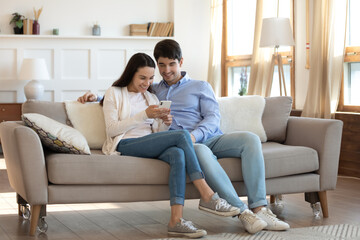 Smiling young caucasian couple sit relax on modern comfortable couch at home using smartphone together, happy man and woman rest on sofa in living room browse internet on cellphone gadget