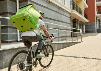 Bearded delivery man in helmet with full thermo bag or backpack looking aside, riding a bike along...