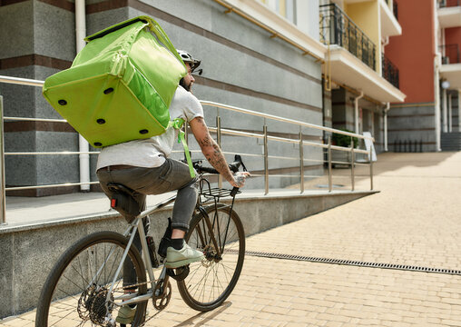 Bearded delivery man in helmet with full thermo bag or backpack looking aside, riding a bike along the city, delivering food. Courier, delivery service concept