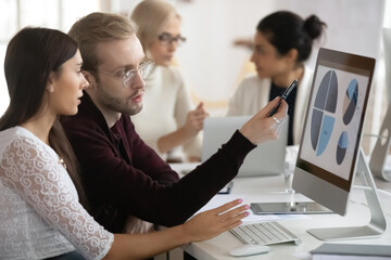 Side view serious young blonde businessman in glasses pointing at graphs on monitor, discussing marketing strategy with young female colleagues. Motivated coworkers working on presentation in office.